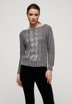 Strickpullover in verschiedenen Grautönen mit einem Kabelmuster, weiter Ausschnitt und gerippten Bündchen. Kombiniert mit schwarzen Hosen, die ein strukturiertes Design zeigen.