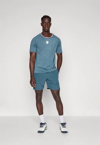 Pánské sportovní oblečení sestávající z tyrkysového trička s krátkými rukávy a kulatým výstřihem s logem, doplněné o odpovídající šortky a bílé tenisky.