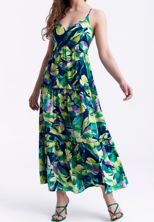 Mujer con un vestido largo sin mangas con estampado tropical multicolor, tirantes finos y sandalias verdes de tacón con tiras.