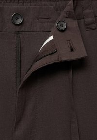 Ceinture de pantalon marron foncé avec deux boutons noirs, fermeture éclair visible et détail de tissu blanc à l'intérieur au niveau de l'ouverture braguette.