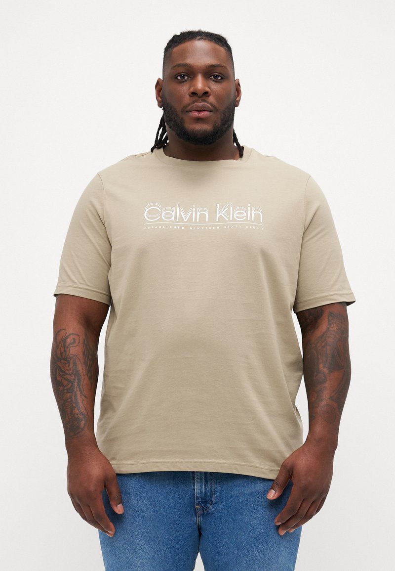 Bež majica kratkih rukava s okruglim izrezom, s bijelim Calvin Klein logotipom ispisanom preko prsnog dijela. Glatka pamučna tekstura.