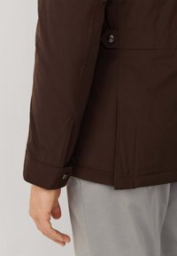 Venstre hånd afslappet ved siden af brun jakkeærme med trykknapsmanchette, og lysegrå bukser mod en ensfarvet baggrund.