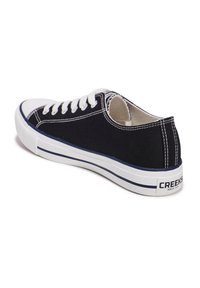 Zapatilla de lona negra con cordones blancos, puntera de goma y suela blanca con un acento azul marino y marca en el talón.