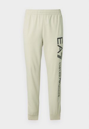 Pantaloni sport crem cu talie elastică, picioare conice și text negru imprimat de-a lungul părții stângi. Textură moale și netedă a materialului.