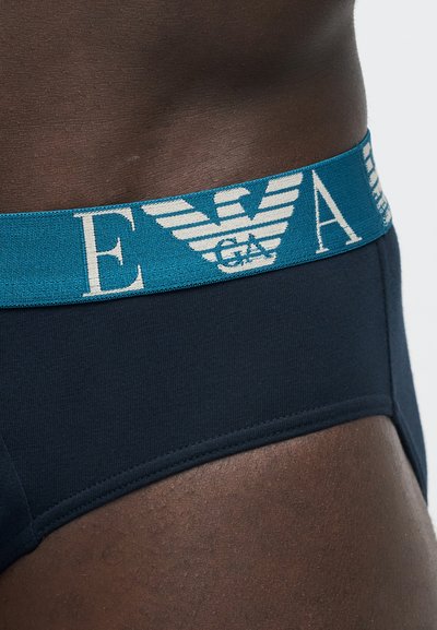 Gros plan sur un slip bleu foncé porté par une personne, avec une taille bleue présentant un logo d'aigle stylisé blanc et des lettres partielles.