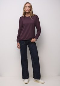 Bordeauxrood top met lange mouwen gecombineerd met donkerblauwe uitlopende spijkerbroek. De spijkerbroek heeft contrasterende stiksels en witte sneakers maken de outfit compleet.