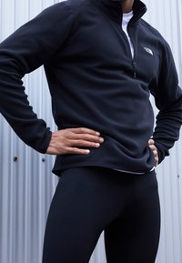 Pullover in pile nero con colletto alto e zip metà, con logo sul petto. Indossato sopra leggings neri, con una texture liscia.