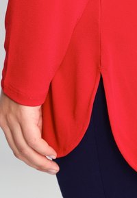 Röd, löst sittande topp med en mjuk yta. Har rundad nederkant och sidsprund. Materialet verkar lätt och ventilerande.