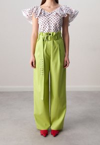 Blusa bianca con motivo floreale viola e maniche arricciate, abbinata a pantaloni a vita alta di un verde chiaro con cintura e dettagli laterali. Scarpe rosse.