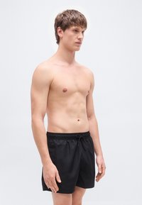 Μαύρο μαγιό βερμούδα από ελαφρύ υλικό, με ελαστική μέση και κορδόνι και άνετη εφαρμογή που φτάνει μέχρι το μέσο του μηρού.