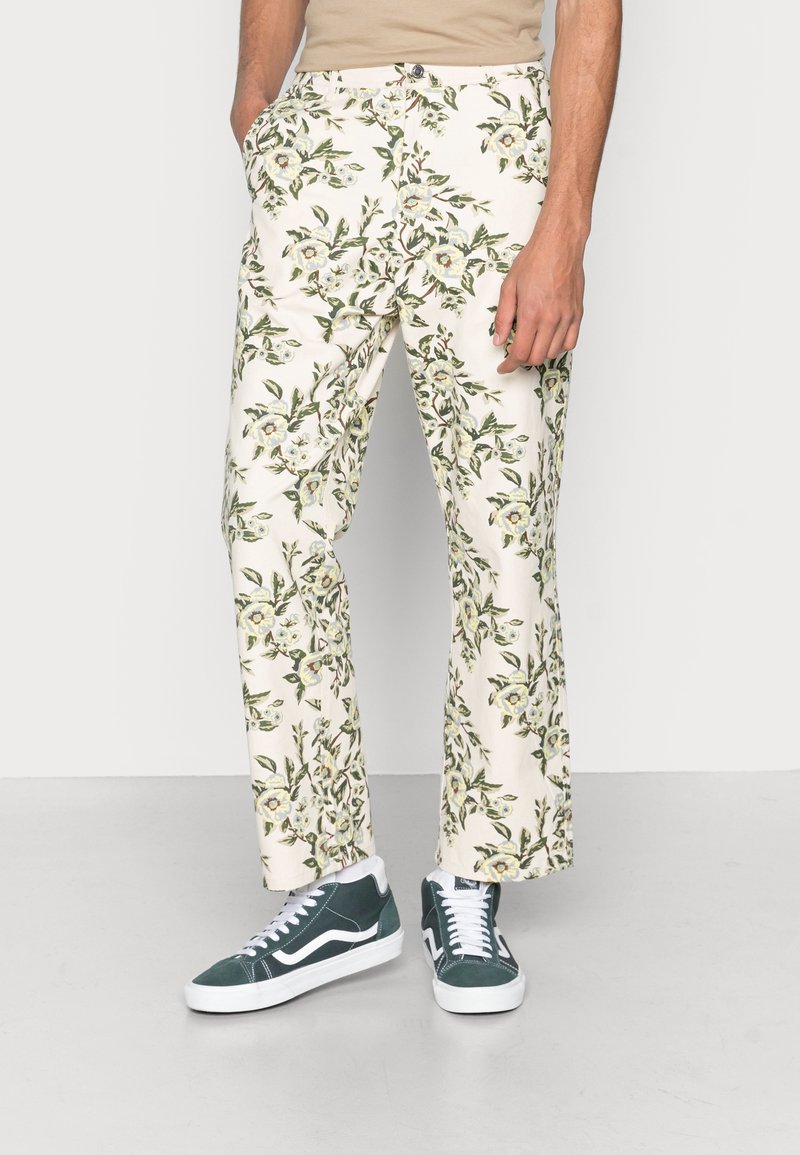Floral-patterned, cream trousers made of lightweight fabric, featuring pockets and a straight-leg design. Worn with green and white sneakers.