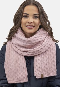 Sciarpa lavorata a maglia di un delicato rosa con un motivo testurizzato, caratterizzata da un bordo a coste e da un piccolo etichetta marrone. Indossata attorno al collo, sovrapposta a un cappotto scuro.
