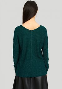 Femme aux longs cheveux bruns portant un pull à manches longues texturé bleu sarcelle et une jupe en cuir noire, vue de dos sur un fond uni.