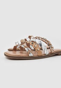Sandales plates avec semelles beige et lanières tressées en argenté, blanc et beige, ornées de clous en métal et de détails en cuir festonné.