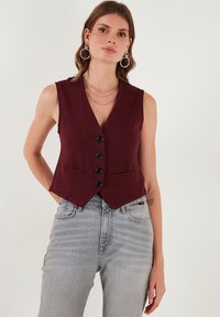 Burgundský bezrukávový sveter s hlbokým V-výstrihom, piatimi čiernymi gombíkmi a dvoma prednými vreckami, skombinovaný s svetlo šedými džínsami s vysokým pásom.