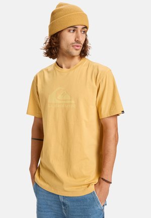 Camiseta de algodón amarilla de manga corta con un sutil estampado de logo, combinada con pantalones cortos de mezclilla y un gorro amarillo acanalado.