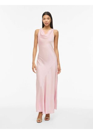 Kvinne står, iført en ermeløs lyserosa satengkjole med drapert utringning og gullflettede åpne hæler på hvit bakgrunn.