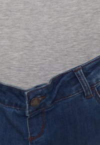 Jeans de ganga em azul escuro com fecho de botão padrão e costura laranja em contraste, combinados com uma t-shirt cinza por baixo.