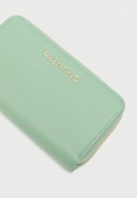 Mētas zaļas ādas maks ar gludu tekstūru, uz kura ir zelta "VALENTINO" zīmogs un aploka rāvējslēdzēja aizdare.