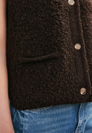 Gilet texturé de couleur brun foncé en matériau duveteux, avec des boutons beiges et une petite poche sur le devant, porté sur un jean en denim bleu.