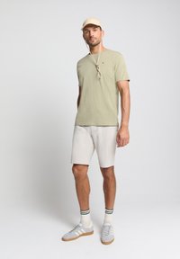 Licht olijfgroene katoenen t-shirt, beige pet, grijze shorts, grijze sneakers met witte accenten en gestreepte sokken. Eenvoudig, casual outfitontwerp.