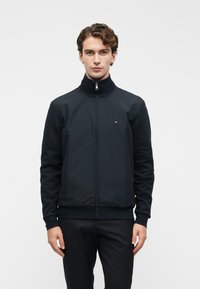 Ανδρας με κυματιστά μαλλιά που φοράει ένα μαύρο μπουφάν με φερμουάρ και ένα μικρό λογότυπο στο στήθος, στέκεται μπροστά σε έναν απλό λευκό φόντο.