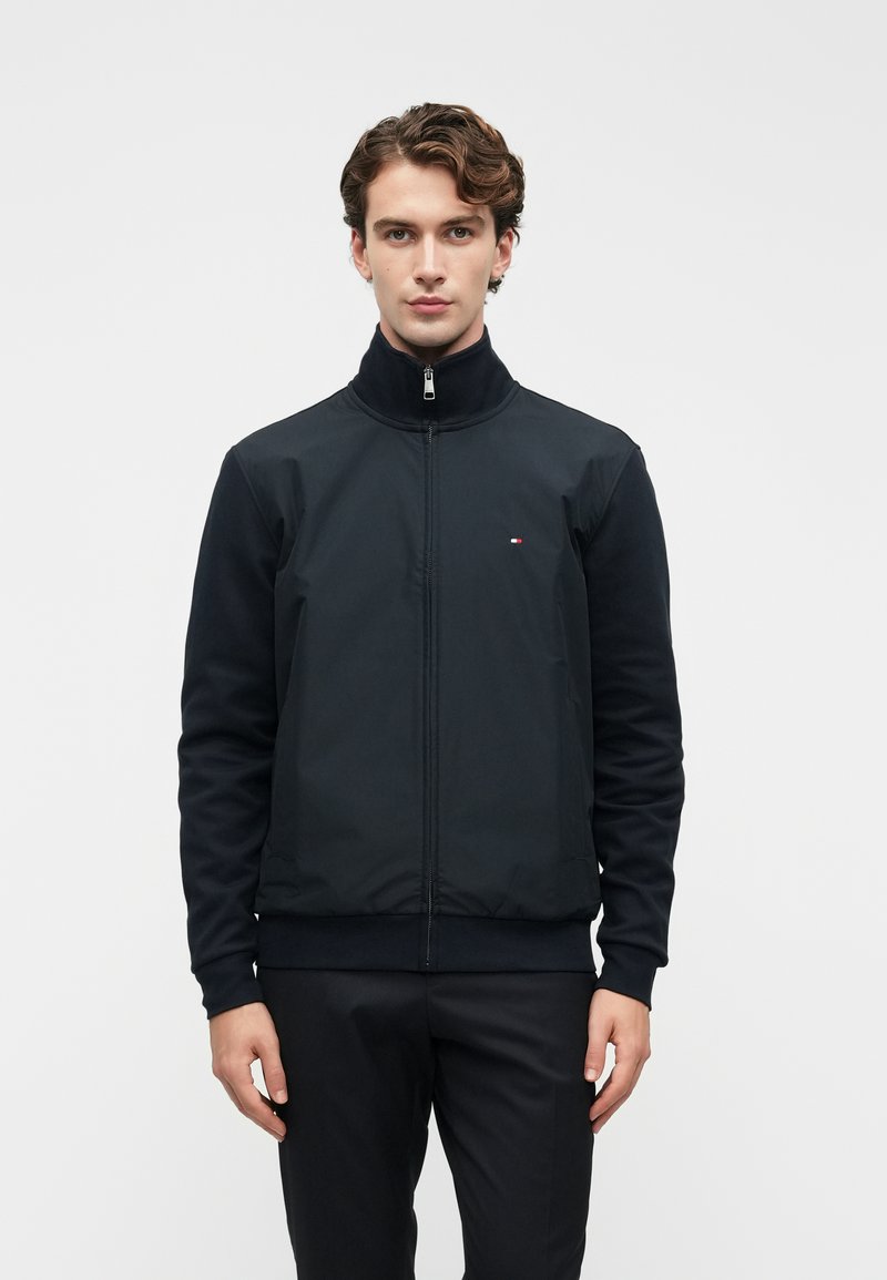 Ανδρας με κυματιστά μαλλιά που φοράει ένα μαύρο μπουφάν με φερμουάρ και ένα μικρό λογότυπο στο στήθος, στέκεται μπροστά σε έναν απλό λευκό φόντο.