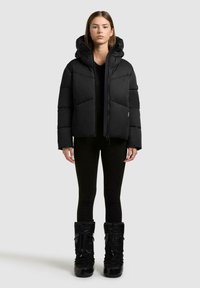 Ung kvinde iført en sort pufferjakke med hætte, sorte leggings og sorte vinterstøvler, stående mod en ensfarvet lys baggrund.