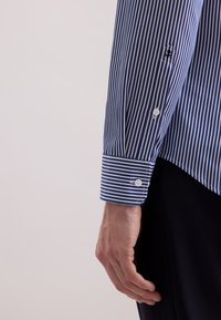 Una mano maschile accanto a una manica di camicia a righe verticali blu e bianche con polsino abbottonato, su uno sfondo chiaro uniforme.
