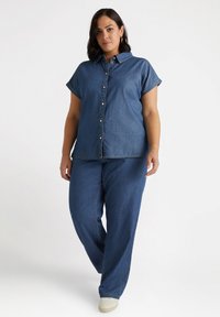 Camisa de denim de mangas cortas, con cierre de botones y cuello, combinada con pantalones de denim holgados y calzado de color claro.
