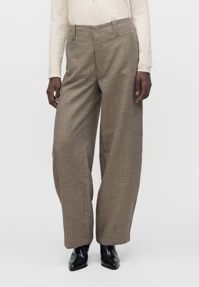 Personne portant un pantalon marron à carreaux taille haute et jambes larges, une chemise crème à manches longues et des bottines noires à bout pointu.