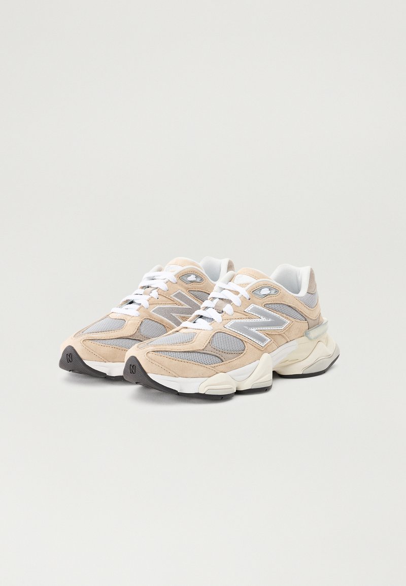 Beige und graue Sneakers aus Mesh und Wildleder, mit einer klobigen weißen Sohle, flachen Schnürsenkeln und einem auffälligen Logo an der Seite.