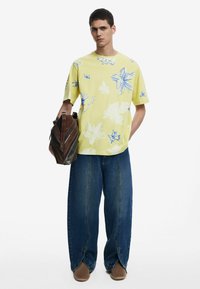 Camisa de manga corta de estampado floral amarillo, combinada con jeans azules holgados y un bolso de cuero marrón. Presenta diseños de flores azules audaces.