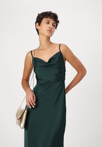 Satin kvällsklänning i mörkgrön, med en cowl neck-design, spaghettiband och en figurnära siluett, i kombination med en guldhandväska.
