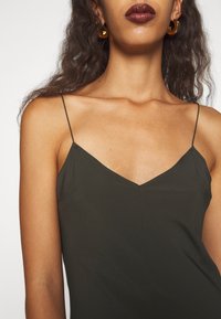 Robe vert olive foncé avec des bretelles spaghetti ajustables et un décolleté en V. Texture lisse avec des coutures minimales et une silhouette cintrée.