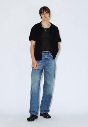 Black short-sleeve shirt over a dark tank top, paired with blue wide-leg jeans and black boots. Silver jewelry accentuates the outfit.