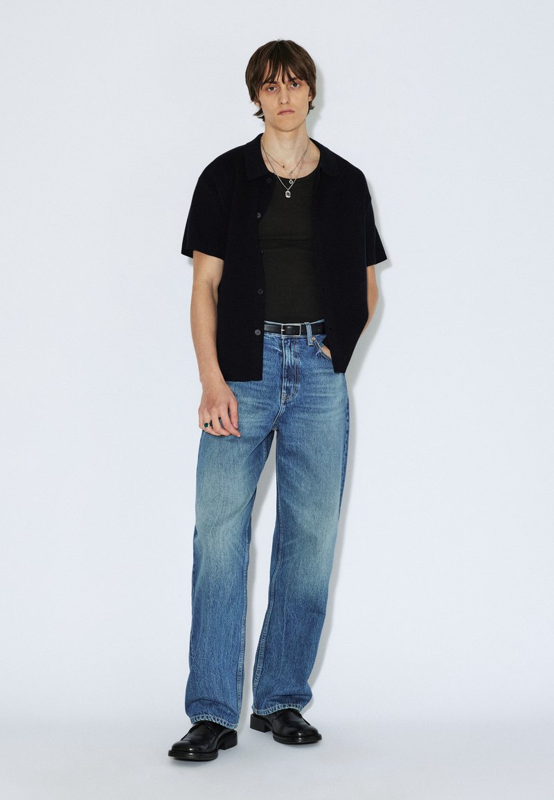 Schwarzes kurzärmeliges Hemd über einem dunklen Tanktop, kombiniert mit blauen, weit geschnittenen Jeans und schwarzen Stiefeln. Silberner Schmuck betont das Outfit.