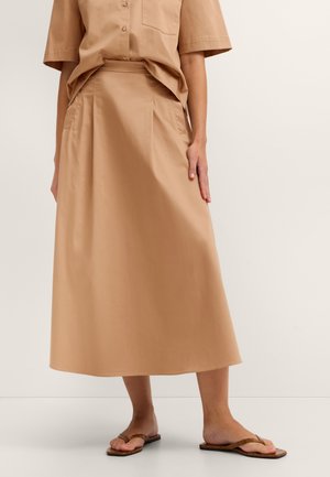 Frau trägt ein beiges kurzärmeliges Hemd mit Knöpfen, das in einen passenden A-Linien-Midirock mit Taschen gesteckt ist, kombiniert mit braunen flachen Sandalen.