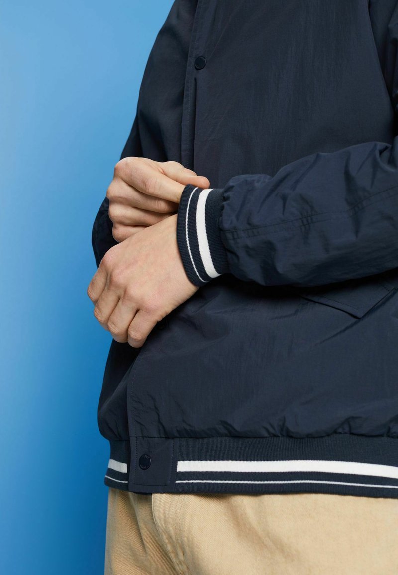Veste bomber bleu marine avec des poignets et une taille à rayures blanches, dotée d'une fermeture à pression et d'un tissu léger. Mains positionnées sur les hanches.