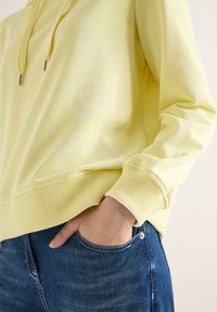 Personne portant un sweat à capuche jaune pâle avec un motif subtil et un jean bleu, la main posée négligemment dans la poche avant.