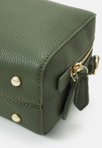 Zelený texturovaný kožený roh tašky se zlatým zipem, dvěma zipsovými táhly a zlatými kovovými nýty na bílém pozadí.