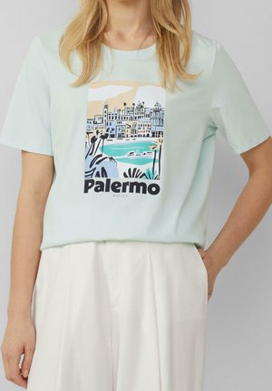 Kobieta w jasnoniebieskiej koszulce z pastelowym widokiem miasta Palermo na Sycylii i palmami, w połączeniu z białymi spodniami.