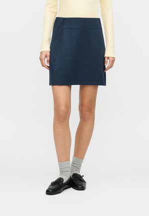 Woman wearing a navy A-line mini skirt, pale yellow long-sleeve top, gray ankle socks, and black loafers standing against a white background.
