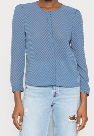 Femme portant un chemisier bleu à manches longues avec un motif chevron, associé à un jean bleu clair déchiré, debout, les bras relâchés le long du corps.