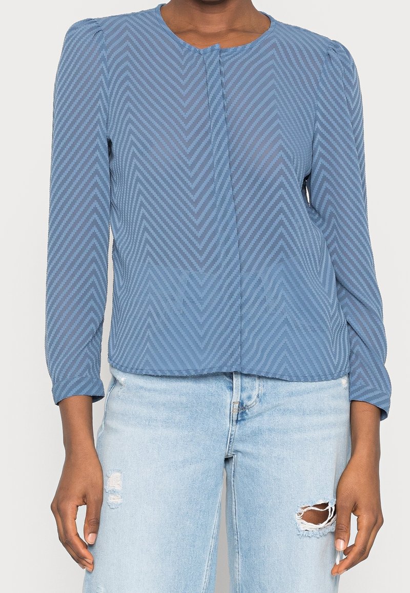 Femme portant un chemisier bleu à manches longues avec un motif chevron, associé à un jean bleu clair déchiré, debout, les bras relâchés le long du corps.