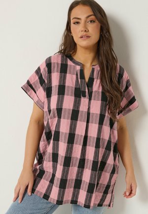 Frau mit langen braunen Haaren, die ein lockeres, rosa-schwarz kariertes kurzärmeliges Oberteil und hellblaue Jeans trägt, vor einer einfarbigen Wand.