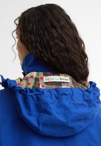 Γυναίκα με καστανά σγουρά μαλλιά που φοράει ένα μπλε μπουφάν με κουκούλα με σχέδιο και ετικέτα «Paul Smith loves Barbour».