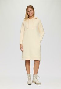 Kapuzenkleid aus leichtem Pfirsichfarbton mit langen Ärmeln und gerippten Bündchen. Kombiniert mit creamfarbenen Schnürstiefeln und einer lässigen Passform.
