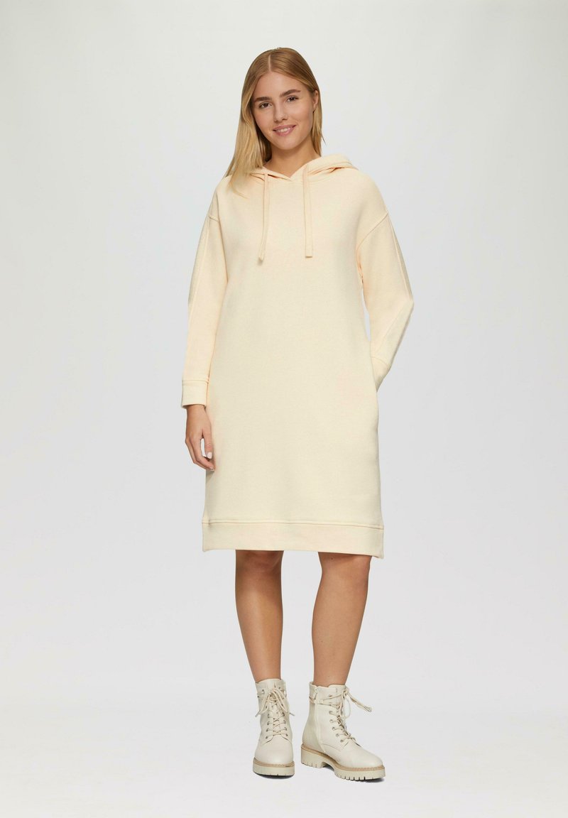 Kapuzenkleid aus leichtem Pfirsichfarbton mit langen Ärmeln und gerippten Bündchen. Kombiniert mit creamfarbenen Schnürstiefeln und einer lässigen Passform.