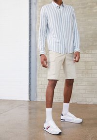 Camicia a righe blu chiaro e bianca con bottoni, pantaloni cargo beige e sneaker bianche con dettagli blu marino e strisce rosse. Tessuto morbido.